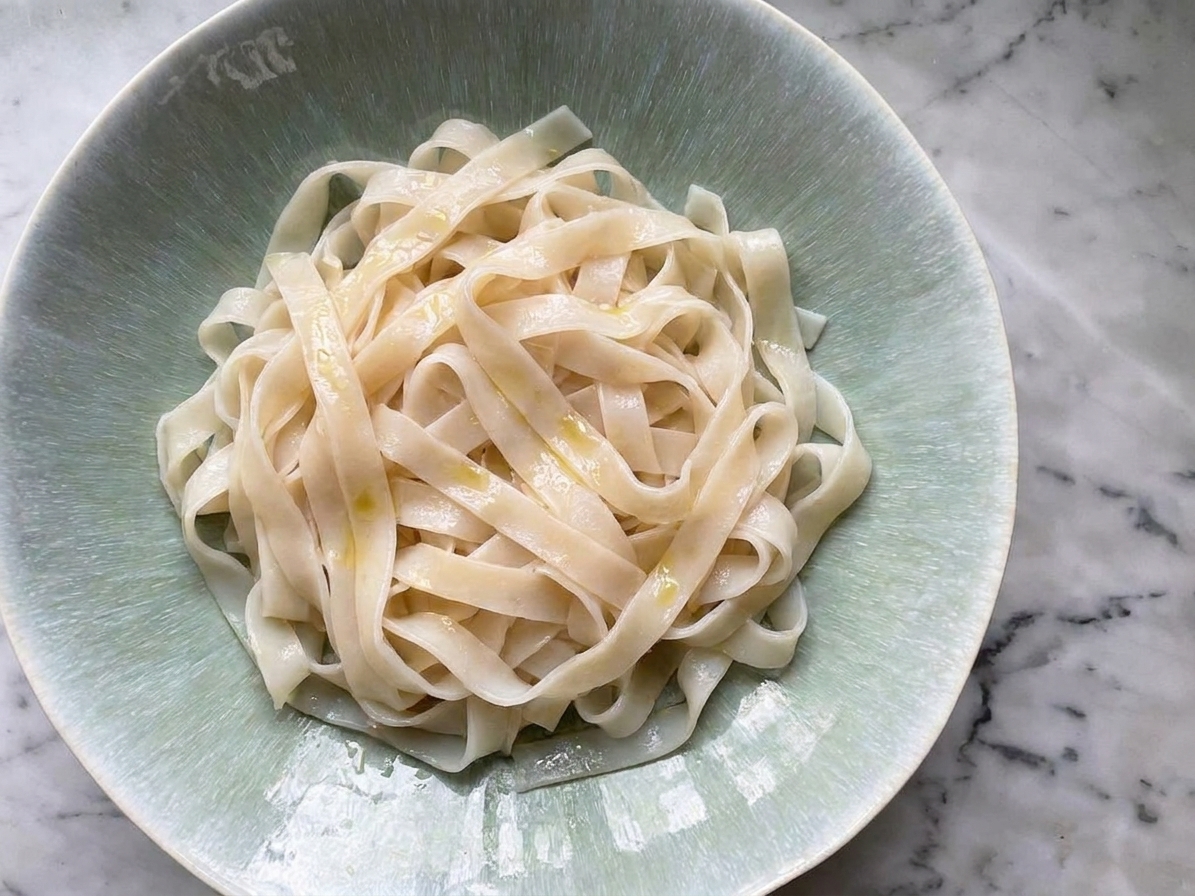 Wide rice noodles loosened on a plate with a light sheen of oil on a kitchen counter