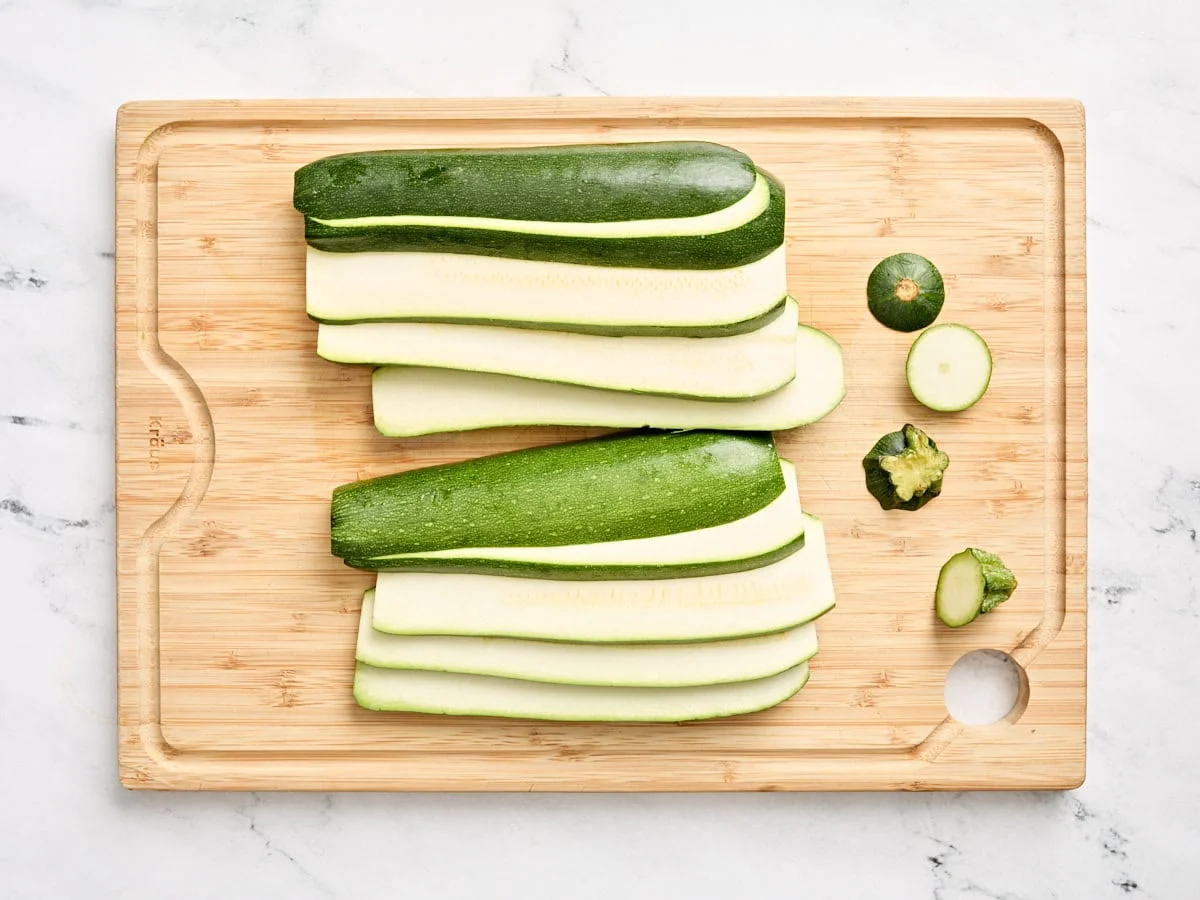 Zucchini cut into thick planks on a wooden cutting board with a chef’s knife beside it on a bright kitchen counter