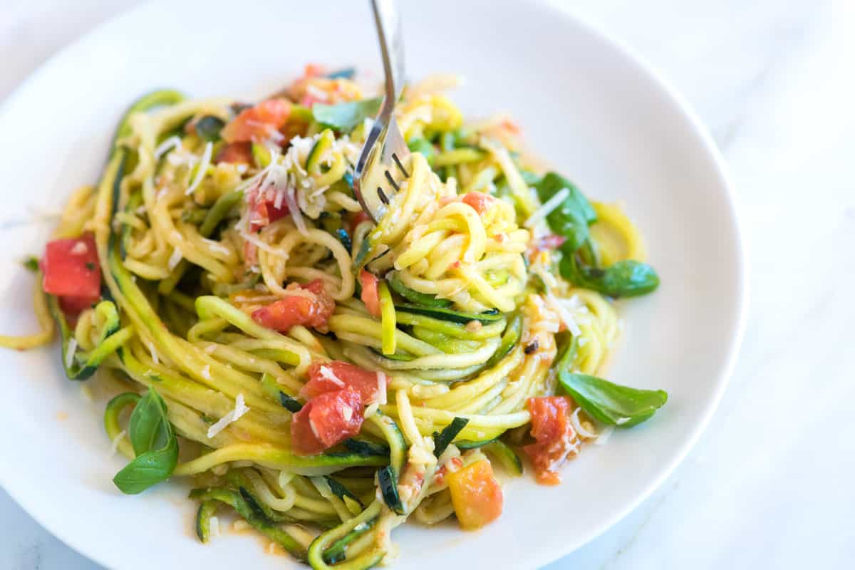 Zucchini noodles being tossed with tongs in a wide skillet on a stovetop with garlic and red pepper flakes
